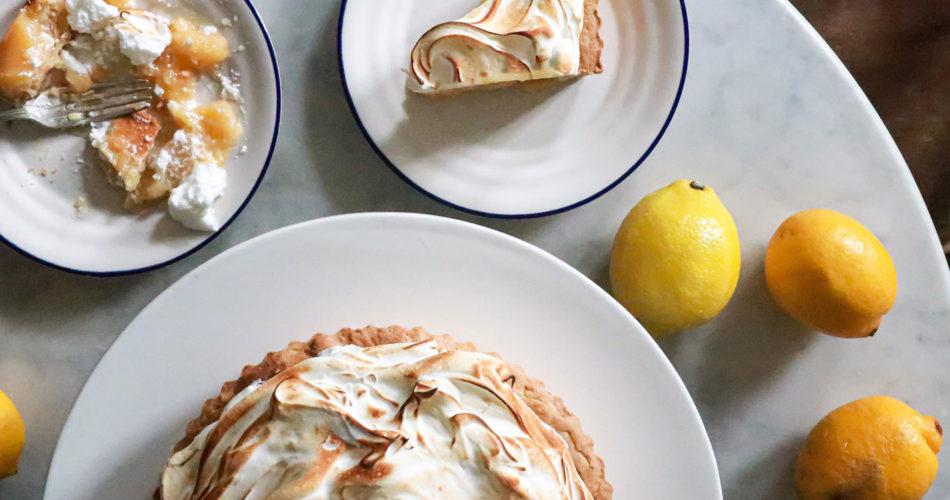 birds eye view of three plates with half a pie topped with meringue on one plate, a slice of the pie on another, and a mushed pie slice on the other, plus three lemons on the table