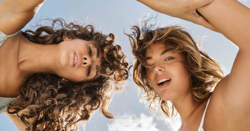 Two women with youthful skin arm in arm looking down at the camera with a sunny blue sky behind them